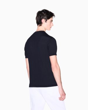 Back view of a man wearing a black short-sleeve knit top with white pants against a white background.