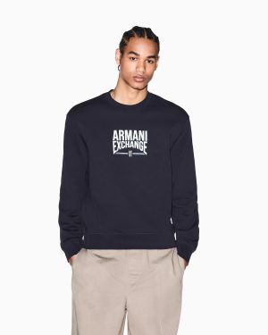 Young man in a navy Armani Exchange sweatshirt with a white logo on the chest, and beige pants, standing against a white background.